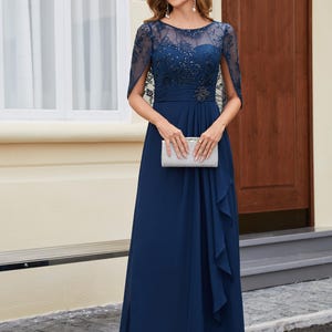 Op de afbeelding: Een marineblauwe formele jurk met een doorschijnend, versierd lijfje en een zwierige rok. De jurk heeft een juwelen riem en een bijpassende clutch. De mouwen zijn doorschijnend en met capes. De vrouw draagt ​​oorhangers en open hakken.