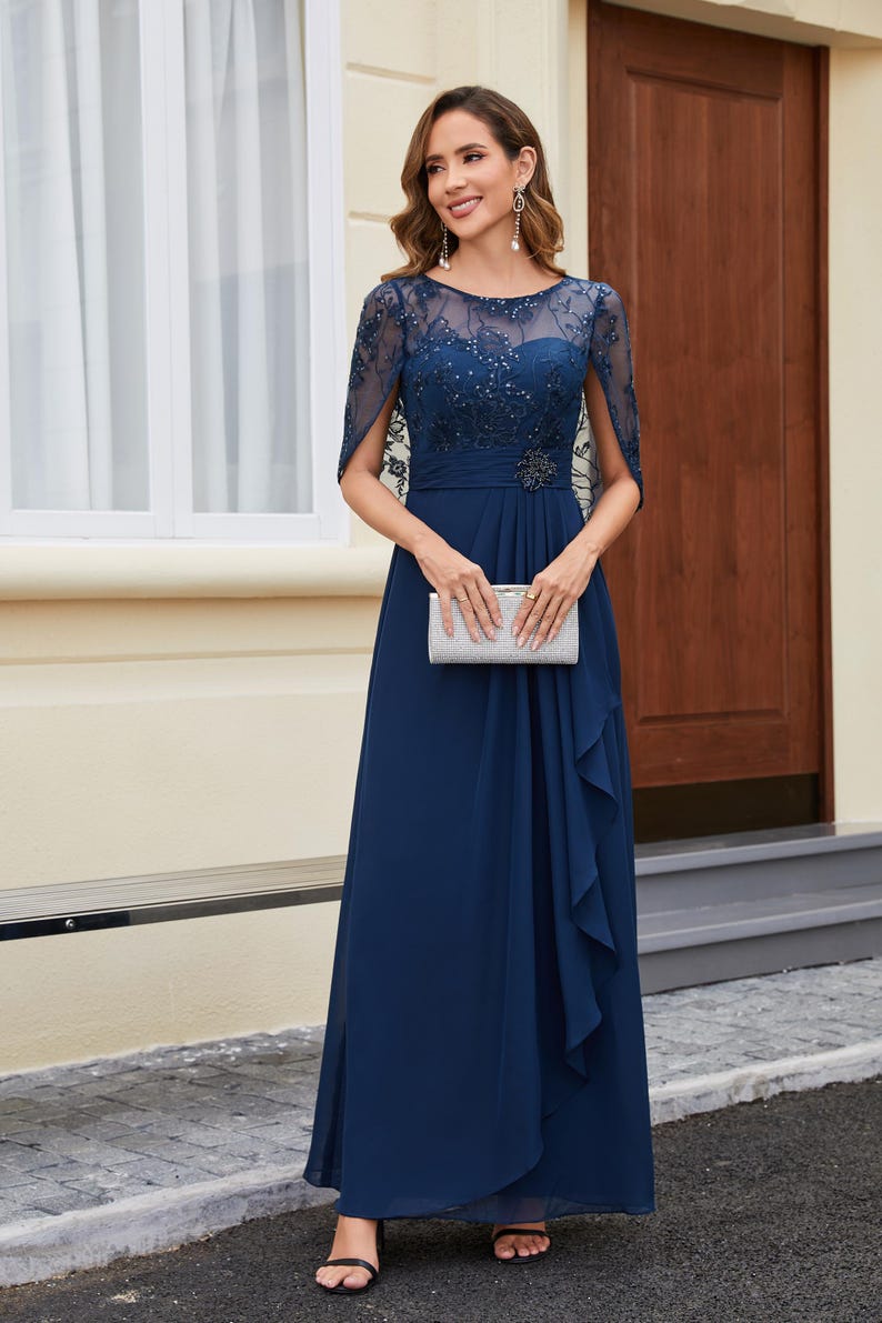 Op de afbeelding: Een marineblauwe formele jurk met een doorschijnend, versierd lijfje en een zwierige rok. De jurk heeft een juwelen riem en een bijpassende clutch. De mouwen zijn doorschijnend en met capes. De vrouw draagt ​​oorhangers en open hakken.