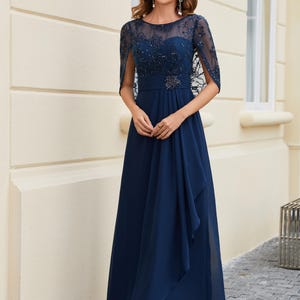 Op de afbeelding: Een marineblauwe formele jurk met een doorschijnende, versierde overlay en capemouwen. De jurk heeft een getailleerd lijfje met een versierde riem en een vloeiende, lange rok. De vrouw draagt ​​sandalen met bandjes.