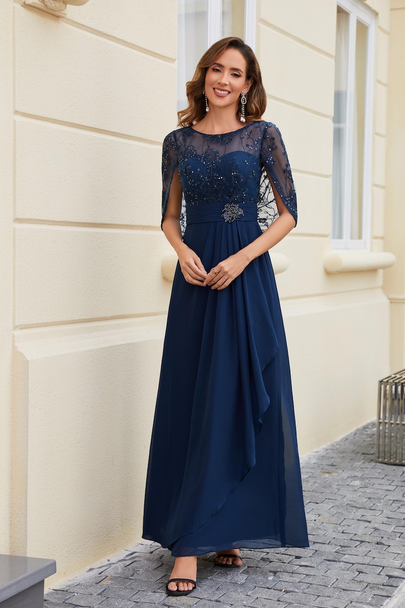 Op de afbeelding: Een marineblauwe formele jurk met een doorschijnende, versierde overlay en capemouwen. De jurk heeft een getailleerd lijfje met een versierde riem en een vloeiende, lange rok. De vrouw draagt ​​sandalen met bandjes.
