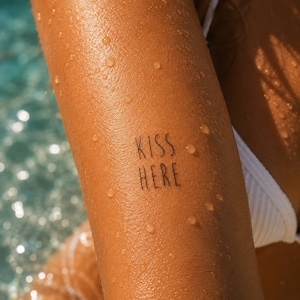 Può includere: Un tatuaggio temporaneo su pelle abbronzata con il testo nero KISS HERE in un semplice carattere sans-serif. La pelle è coperta da piccole gocce d'acqua e una parte di un indumento in tessuto bianco a coste è visibile sul lato destro.
