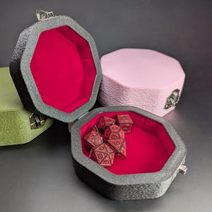 May include: Four octagonal dice boxes in shades of green, pink, and grey, with a red interior. One box is open, revealing a set of red dice. The boxes have a textured exterior and metal clasps.