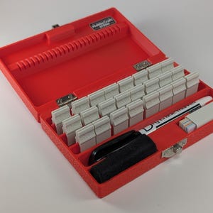 Puede incluir: Caja de plástico roja abierta con exterior texturizado. En el interior hay filas de pequeños sellos de letras grises, un rotulador negro, un rotulador blanco y un pequeño recipiente blanco. La caja está abierta, revelando su contenido.
