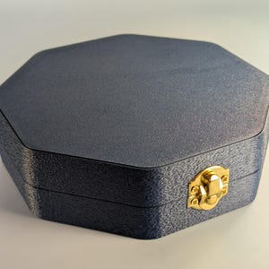 Pu&ograve; includere: Una scatola ottagonale blu scuro con una superficie strutturata. La scatola ha una chiusura color oro sul davanti. La scatola &egrave; chiusa e sembra essere fatta di un materiale robusto.