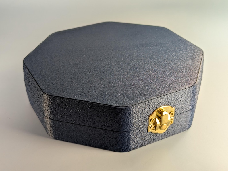 Pu&ograve; includere: Una scatola ottagonale blu scuro con una superficie strutturata. La scatola ha una chiusura color oro sul davanti. La scatola &egrave; chiusa e sembra essere fatta di un materiale robusto.