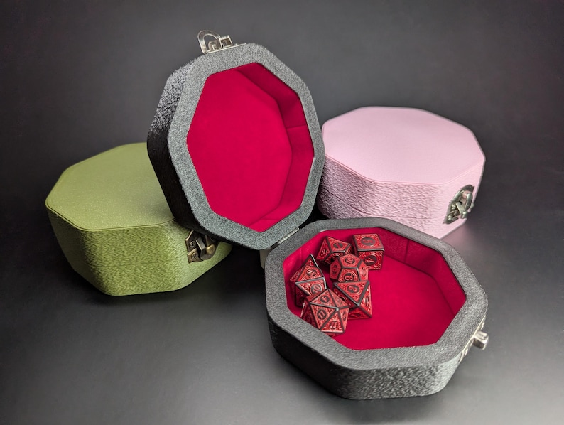 May include: Four octagonal dice boxes in shades of green, pink, and grey, with a red interior. One box is open, revealing a set of red dice. The boxes have a textured exterior and metal clasps.