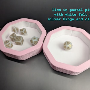Może przedstawiać: Pastelowo-r&oacute;żowe pudełko na kości, o średnicy 11 cm, pokryte filcem, z białym wnętrzem oraz srebrnym zawiasem i zapięciem. Otwarte pudełko prezentuje kilka kości z złotymi oznaczeniami. Tekst na zdjęciu brzmi: "11cm in pastel pink, with white felt and silver hinge and clasp."
