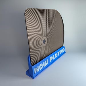 Può includere: Un espositore per dischi in vinile marrone e nero con la scritta "NOW PLAYING" in bianco su una base blu. Il disco presenta un motivo a scanalature circolari.
