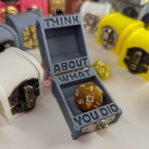 Könnte beinhalten: Eine graue Miniatur-Schatztruhe mit den Worten "THINK ABOUT WHAT YOU DID" eingraviert, geöffnet, um einen braunen und goldenen d20-Würfel zu enthüllen. Andere farbenfrohe Truhen sind im Hintergrund.