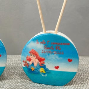 Può includere: Due oggetti decorativi rotondi, blu e bianchi, con bastoncini di legno. Ognuno presenta un'illustrazione di sirena dei cartoni animati, cuori rossi e testo. Per una festa o un evento.