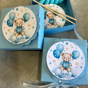 Może przedstawiać: Jasnoniebieskie pudełka na prezenty z grafiką misia trzymającego balony. Miś siedzi na chmurze z imieniem "Noah" poniżej. Pudełka są zawiązane jasnoniebieską wstążką.