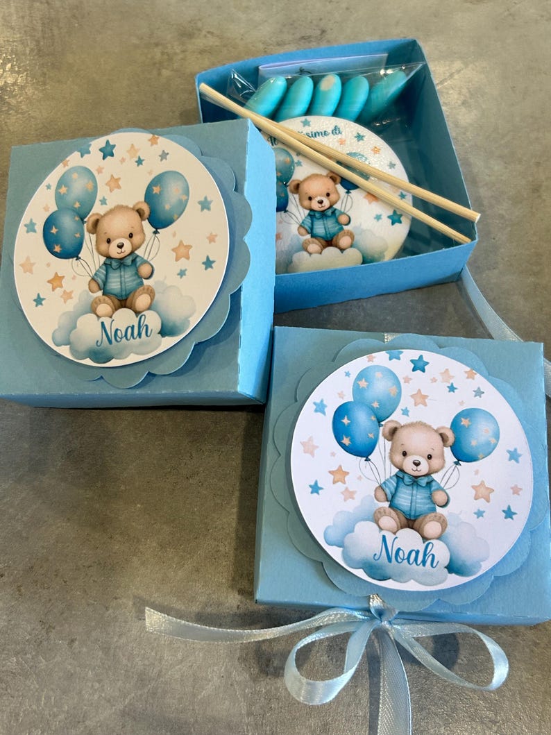 Może przedstawiać: Jasnoniebieskie pudełka na prezenty z grafiką misia trzymającego balony. Miś siedzi na chmurze z imieniem "Noah" poniżej. Pudełka są zawiązane jasnoniebieską wstążką.