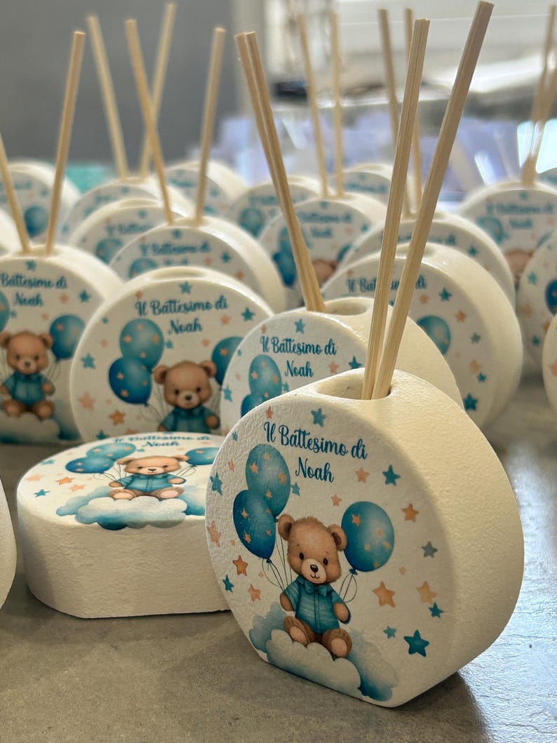 Może przedstawiać: Biały, okrągły ceramiczny odświeżacz powietrza z drewnianymi patyczkami. Projekt przedstawia misia trzymającego niebieskie balony, chmury i gwiazdy. Tekst brzmi "Il Battesimo di Noah". Idealny na baby shower lub chrzciny.