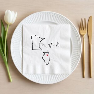 May include: A white paper napkin with a black outline of the states of Minnesota and Illinois, connected by a dotted line. The initials "H + K" are written in cursive. A red heart is in Illinois. The napkin is on a white plate with gold silverware.