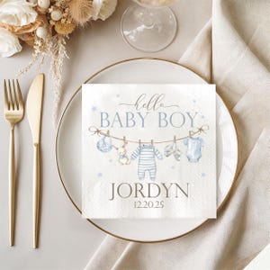 May include: A white paper napkin with a baby shower theme. The napkin features the text "hello BABY BOY" in script, along with illustrations of baby clothes and a bunny. The name "Jordyn" and the date "12.20.25" are also printed on the napkin.