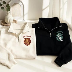 Op de afbeelding: Een wit en een zwart sweatshirt met een korte rits. Het witte sweatshirt heeft een rood en goud Gryffindor-embleem met een leeuw. Het zwarte sweatshirt heeft een groen en zilver Slytherin-embleem met een slang. Beide hebben zilveren ritsen.
