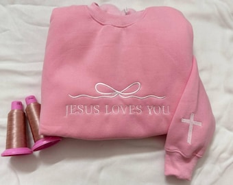 May include: A pink sweatshirt with white embroidered text that reads "JESUS LOVES YOU". The design includes a bow and a cross on the sleeve. Two spools of pink thread are next to the sweatshirt.