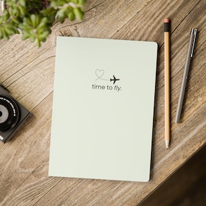 May include: A light green notebook with the text "time to fly" and a small airplane graphic. A pencil and a pen are next to the notebook on a wooden surface. A camera is visible in the corner.