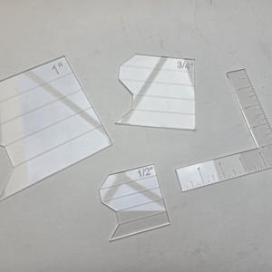 May include: Four clear acrylic quilting templates with measurements. The templates include a 1-inch, 3/4-inch, and 1/2-inch marked pieces, along with a ruler. The quilting tools are designed for precise fabric cutting and sewing.