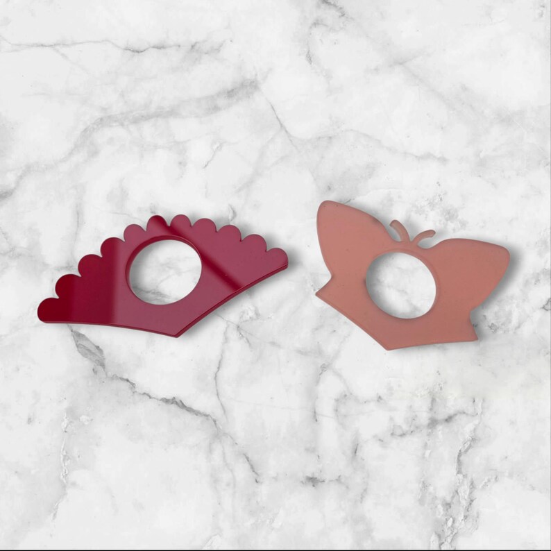 May include: Two decorative napkin rings. One is a deep red fan shape, and the other is a peach-coloured butterfly shape. Both have a circular opening in the centre. The background is a white marble pattern.