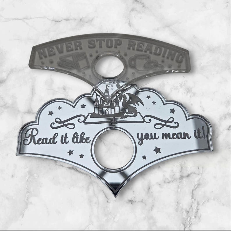 May include: A silver bookmark with two linked parts. The top section reads "NEVER STOP READING" and has book and coffee cup illustrations. The bottom section, cloud-shaped, says "Read it like you mean it!" and features a castle and dragon design.