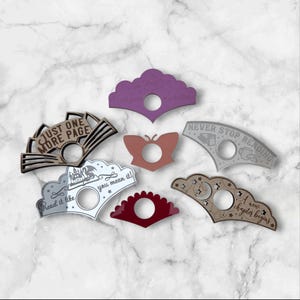 Puede incluir: Una colección de sujetapáginas decorativos en varias formas y colores, incluyendo morado, marrón y plateado. Cada sujetapáginas presenta una abertura circular y textos como "Just One More Page" y "Never Stop Reading".