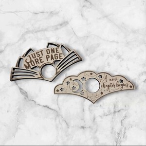 May include: Two wooden book page holders. One is fan-shaped with the text "JUST ONE MORE PAGE". The other is cloud-shaped with stars, moons, and the text "A new chapter begins". Both have a circular hole for the thumb.