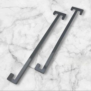 May include: Two dark grey metal shelf brackets with a modern, minimalist design. Each bracket has a long, straight arm with a right-angled hook at each end. The brackets are displayed against a white and grey marble background.