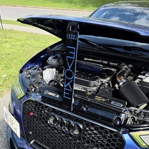 Op de afbeelding: Een zwart verticaal motorruimtepaneel met het Audi-logo en het woord EVOKE in blauwe letters, geïnstalleerd in een blauwe auto met een open motorkap. Het paneel bevat een klein Instagram-label en goudkleurige handgeschreven tekst.