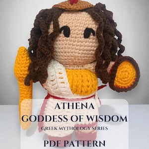 May include: Crocheted Athena doll from the Greek Mythology Series. The doll has brown curly hair, a tan face, and is wearing a yellow and white outfit. The doll is holding a yellow staff and a shield. Text on the image reads "ATHENA GODDESS OF WISDOM PDF PATTERN".