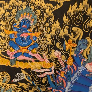 May include: A colorful Tibetan Buddhist thangka painting depicting a deity with multiple arms and hands, surrounded by intricate patterns and symbols. The deity is blue and has a fierce expression, holding a musical instrument in one hand. The background is black with gold and red accents.