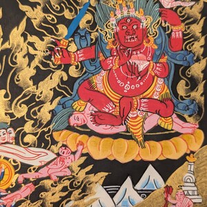 May include: A colorful Tibetan Buddhist painting depicting a deity with a red body, multiple arms, and a fierce expression. The deity is seated on a lotus throne surrounded by clouds and other figures. The painting is done in a traditional style with gold accents.