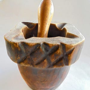 Op de afbeelding: Een houten vijzel en stamper set. De vijzel is donkerbruin met een uitgesneden ruitpatroon rond de rand. De stamper is lichtbruin en glad en afgerond.