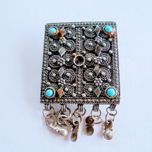 May include: A vintage silver-toned rectangular brooch with intricate filigree detailing, small turquoise-colored stones, and dangling charms. The brooch features a detailed pattern of swirls, diamonds, and circular elements.