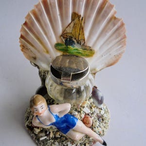 Op de afbeelding: Een vintage inktpot in de vorm van een zeeschelp met een geschilderde zeilbootscène. Een kleine glazen inktpot met een zilveren deksel staat voor de schelp. Een porseleinen beeldje van een vrouw in een blauw badpak ligt op een bed van zand en schelpen.