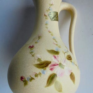 Peut inclure: Pichet en céramique crème avec une anse et un motif floral. Le pichet présente un motif peint de fleurs roses et blanches, de feuilles vertes et d'accents bleus. Une étiquette est apposée sur le pichet.