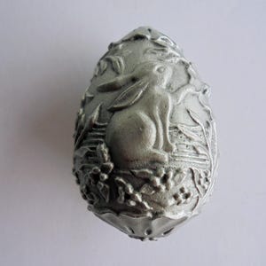 Può includere: Un ornamento argentato a forma di uovo con un rilievo dettagliato di un coniglio circondato da foglie. L'oggetto a tema pasquale ha una superficie strutturata e un fondo arrotondato.