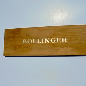 May include: A rectangular wooden box with the word "BOLLINGER" printed in gold lettering. The box is a light brown colour and appears to be made of wood. The background is a light blue colour.