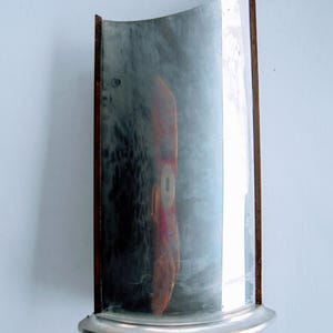 Könnte beinhalten: Ein gebogener, antiker Spiegel mit silbernem Sockel und Holzrahmen. Die Oberfläche des Spiegels reflektiert ein verschwommenes Bild, und der Rahmen hat eine dunkelbraune Oberfläche. Der Spiegel ist an einer hellblauen Wand befestigt.