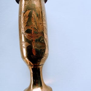 Puede incluir: Un jarrón vintage de color dorado, con una base acampanada y un cuerpo delgado y alargado. El jarrón presenta diseños de hojas grabadas y una parte superior ligeramente más ancha. La superficie metálica refleja la luz, creando reflejos y sombras.