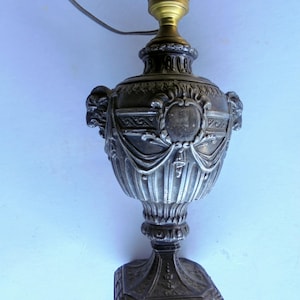 May include: An antique silver-toned table lamp with ornate detailing. The lamp features a decorative urn-shaped body with relief carvings, a square base, and a brass lamp holder. The lamp is designed with a cord for electrical use.