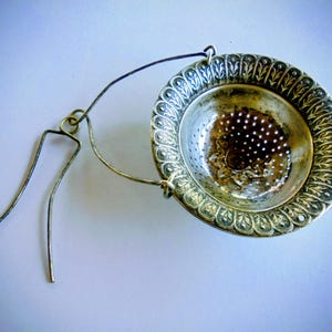 Op de afbeelding: Een theezeef van zilverkleurig metaal met een ronde kom, een geperforeerde bodem en een versierde rand met bladmotief. De zeef is voorzien van een dunne, gebogen draadgreep en een apart metalen steunhulpstuk met twee tanden.