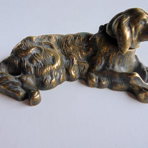 Pode incluir: Uma estatueta de cão de bronze em posição reclinada. A escultura tem uma textura de pelo detalhada e é fundida em um tom metálico quente. A cabeça do cão está virada para o lado e suas patas estão estendidas. Um item decorativo.