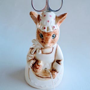 Può includere: Un portafili in ceramica a forma di coniglietto. Il coniglietto indossa un vestito bianco e un cappello a pois rosa. Le forbici sono argentate e sono inserite nella testa del coniglietto. Un filo bianco pende dalle mani del coniglietto.
