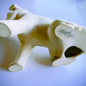 Pode incluir: Estatueta de c&atilde;o em cer&acirc;mica creme com superf&iacute;cie texturizada. O c&atilde;o est&aacute; posando com as patas dianteiras levantadas e a cabe&ccedil;a inclinada. A parte inferior do c&atilde;o tem um buraco e escrita.