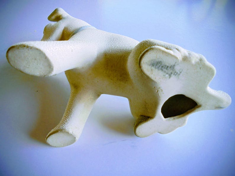 Pode incluir: Estatueta de c&atilde;o em cer&acirc;mica creme com superf&iacute;cie texturizada. O c&atilde;o est&aacute; posando com as patas dianteiras levantadas e a cabe&ccedil;a inclinada. A parte inferior do c&atilde;o tem um buraco e escrita.