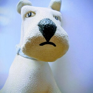 Pode incluir: Estatueta de c&atilde;o em cer&acirc;mica creme com superf&iacute;cie texturizada. O c&atilde;o tem nariz preto, olhos azuis e coleira azul. As orelhas do c&atilde;o est&atilde;o em p&eacute; e o design geral &eacute; simples e charmoso.
