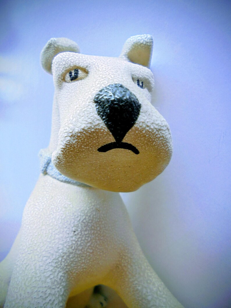 Pode incluir: Estatueta de c&atilde;o em cer&acirc;mica creme com superf&iacute;cie texturizada. O c&atilde;o tem nariz preto, olhos azuis e coleira azul. As orelhas do c&atilde;o est&atilde;o em p&eacute; e o design geral &eacute; simples e charmoso.