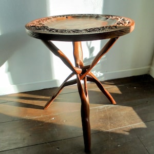 Op de afbeelding: Een handgemaakte houten bijzettafel met een rond blad en drie poten. Het tafelblad heeft ingewikkelde gesneden bloemenmotieven langs de rand. De tafel is gemaakt van donkerbruin hout en staat op een houten vloer.