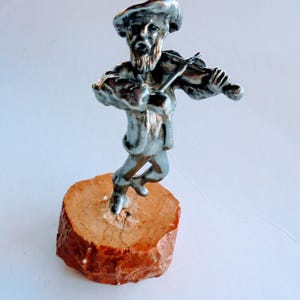 Könnte beinhalten: Silberfarbene Figur eines Geigers auf einem kleinen, runden, braunen Holzsockel. Die Figur ist detailliert mit Bart und Hut. Der Sockel hat einen Durchmesser von etwa 5 cm.